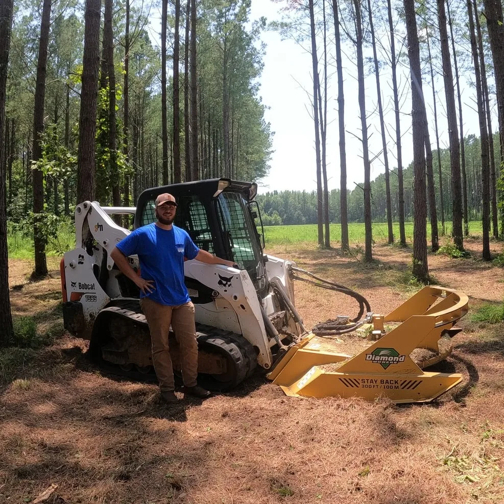 Baylor Weathers leaning on a Bobcat skid steer fitted with a mulching head, in a stand of pines.